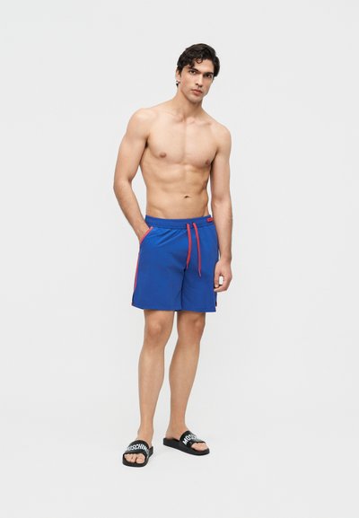 Skjorteløs mann iført blå badeshorts med rød snor og svarte Moschino slidesandaler, stående med den ene hånden i lommen.