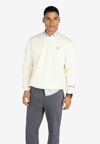 Sudadera color crema con cuello redondo, que presenta un pequeño logo negro en el pecho y un detalle de texto en la manga, usada sobre una camisa azul.