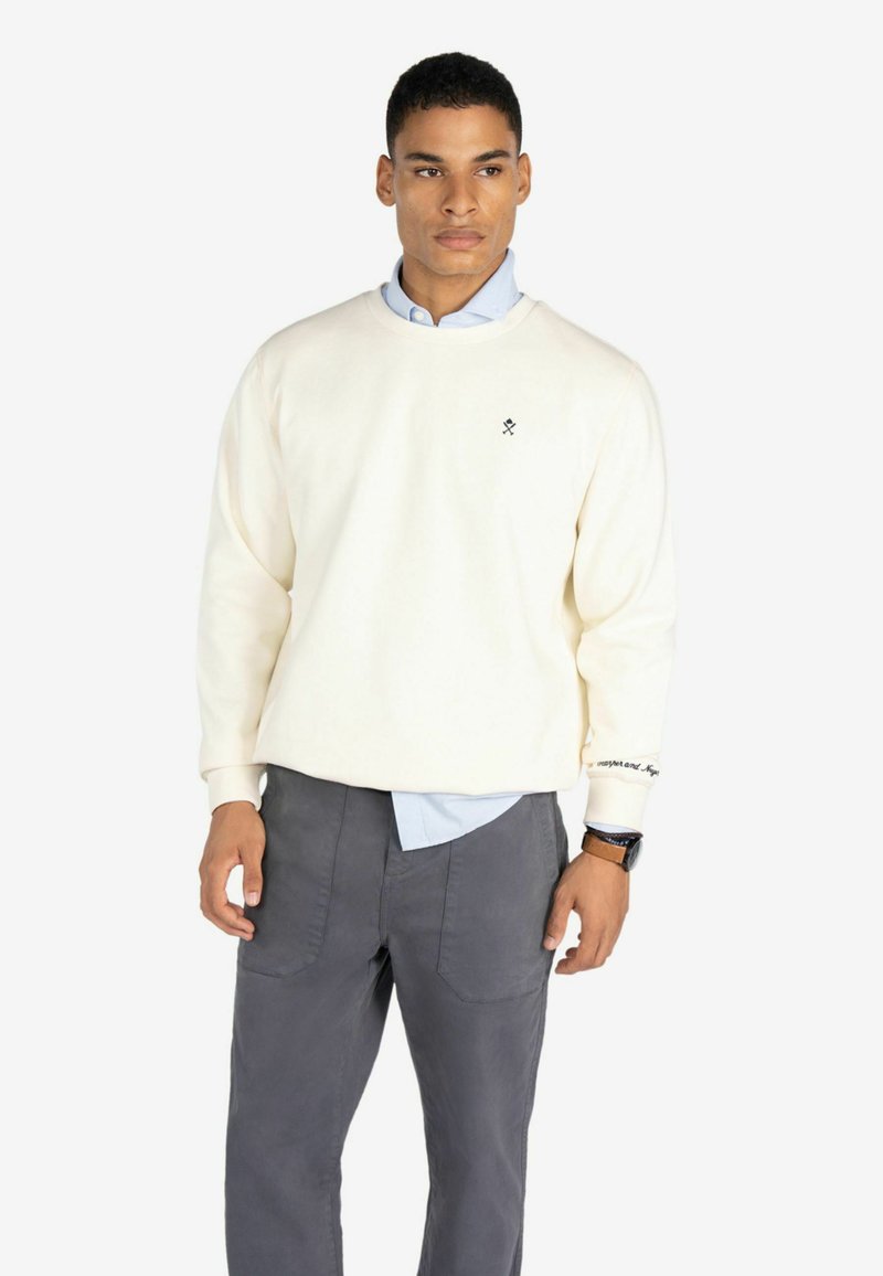 Sudadera color crema con cuello redondo, que presenta un pequeño logo negro en el pecho y un detalle de texto en la manga, usada sobre una camisa azul.