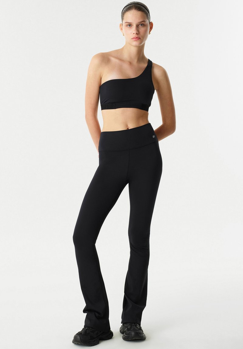 Zwarte sportbeha met één schouderband, gecombineerd met hoge zwarte flare leggings, gemaakt van rekbaar materiaal en met een gladde textuur.