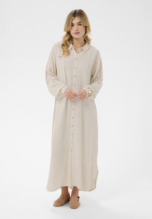 Žena s vlnitými blond vlasmi, oblečená v dlhých béžových šatách na gombíky a s tkanými opálenými balerínami, stojaca so založenými rukami na bielom pozadí.