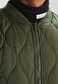 Pikowana zielona kurtka bombowiec z kołnierzem stójką, zamkiem błyskawicznym i teksturowanym wzorem z gładkimi akcentami materiału.