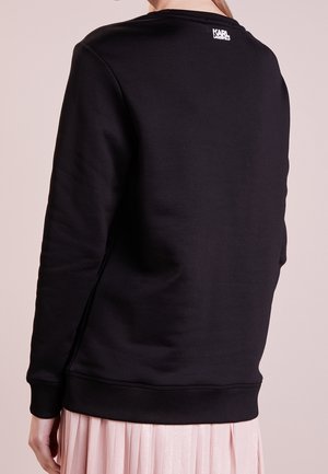 Personne portant un sweat-shirt noir à manches longues avec le logo Karl Lagerfeld sur le haut du dos, associé à une jupe plissée rose clair.