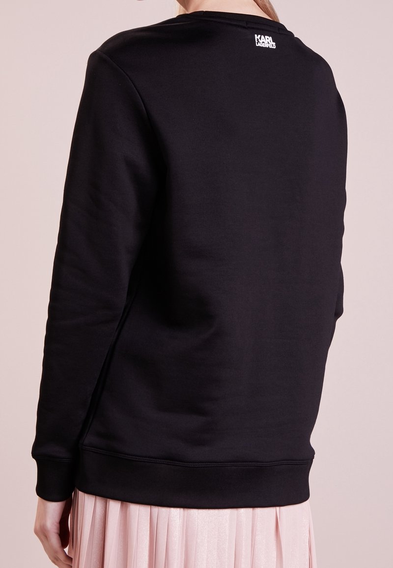 Personne portant un sweat-shirt noir à manches longues avec le logo Karl Lagerfeld sur le haut du dos, associé à une jupe plissée rose clair.