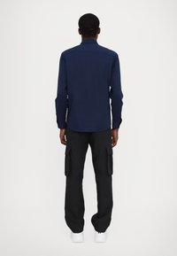 Camicia a maniche lunghe blu navy con colletto e polsini con bottoni, abbinata a pantaloni cargo neri con tasche laterali e indossata con scarpe da ginnastica bianche.