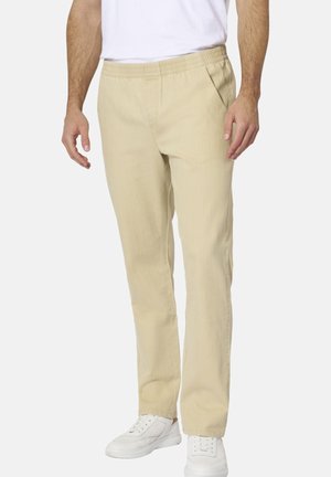 Mann trägt beige elastische Bund gerade geschnittene Hose, weiße Sneaker und ein weißes Hemd und steht vor einem einfarbigen Hintergrund.