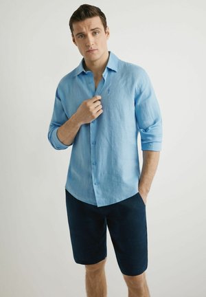 Młody mężczyzna poprawia jasnoniebieską koszulę z długim rękawem, ubrany w granatowe, sięgające kolan szorty, stoi na jednolitym białym tle.