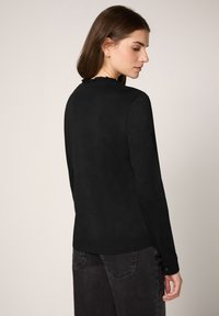 Czarna bluzka z długim rękawem, z falbanką przy dekolcie i dopasowanym kroju, wykonana z miękkiego materiału, zestawiona z ciemnymi dżinsami.