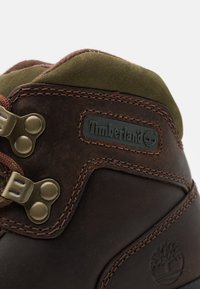 Rudos odinės botai su žaliu zomšos apvadu, papuošti metaliniais akcentais, siuvinėtais raštais ir Timberland logotipu. Matomas tekstūruotas paviršius ir dizaino detalės.
