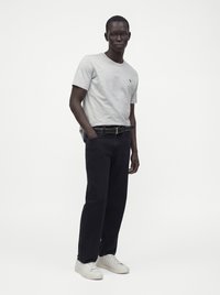 Jeune homme debout, une main dans la poche, portant un t-shirt gris clair, un pantalon noir avec ceinture et des baskets blanches sur un fond uni.