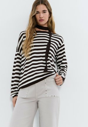 Femme aux cheveux longs et ondulés portant un haut à manches longues rayé noir et blanc, une écharpe noire autour du cou et un pantalon gris clair avec des clous en métal.