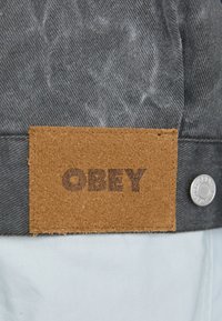 Gray fabric with a textured surface features a brown leather label embossed with "OBEY" and a metal snap fastener on the side.