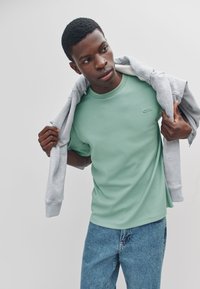 Mintgroen T-shirt met korte mouwen en een ronde halslijn, voorzien van een klein grijs logo op de borst; gecombineerd met lichtblauwe denim jeans.