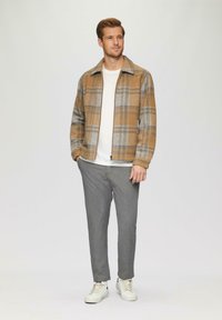 Bruine en grijze geruite jas met een rits aan de voorkant, gedragen over een wit shirt, gecombineerd met grijze broek en witte sneakers. Soepele stoftextuur.