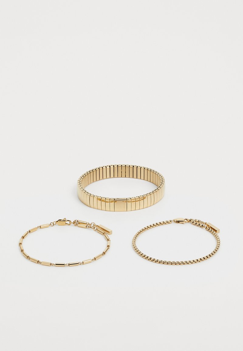 Tre gullarmbånd: et fleksibelt armbånd med vertikale riller, en kjedelink med forlengede segmenter, og en tynn, glatt kjede med små akenter.