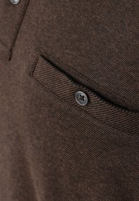 Braun strukturiertes Material mit einer vorderen Tasche, die einen silbernen Knopf aufweist. Das Material wirkt strapazierfähig und hat ein gewebtes Muster.