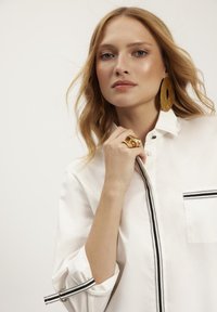 Biała koszula z kieszonką z przodu i czarnym zamkiem błyskawicznym. Modelka nosi duże złote kolczyki i masywny złoty pierścionek. Gładki, lekki materiał.