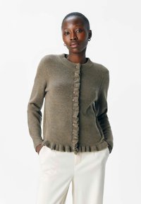 Olivegrøn cardigan lavet af blødt stof, med rund halsudskæring, knapper foran og flæsekanter. Sat sammen med lyse bukser.