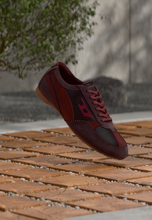Sneaker marrone scuro e nero con lacci rossi e logo rosa che fluttua sopra una superficie pavimentata in legno con piccole pietre bianche tra le assi.
