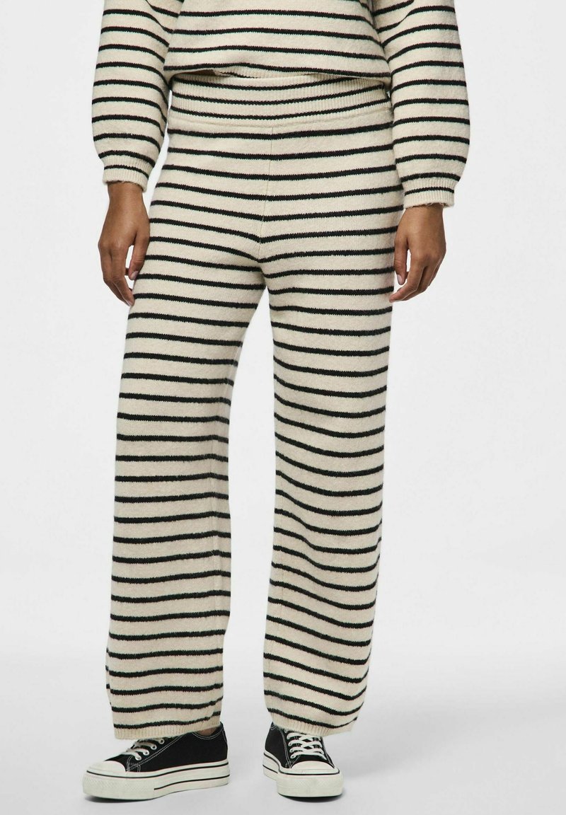 Riba-lõngaga mugavusriiete komplekt kreemjas värvitoonis, mustade ja roheliste triipudega, sisaldab laia säärega pükse ja lühikest sviitrit. Hästi nähtavad sokid ja tossud.