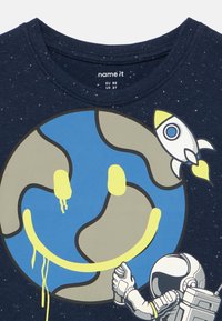 Astronaut malujący sprayem żółtą buźkę na Ziemi, z lecącą rakietą w pobliżu na ciemnoniebieskim tle koszulki.