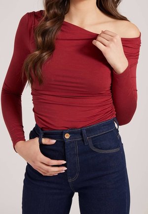 Kvinde iført en langærmet, skulderfri vinrød bluse og højtaljede mørkeblå jeans, med den ene hånd nær kravebenet og den anden i lommen.