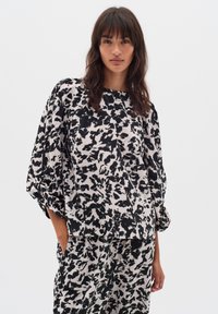 Femeie îmbrăcată cu un top alb-negru, cu un model abstract, și pantaloni asortați, având mâinile în buzunare, stând în fața unui fundal alb simplu.