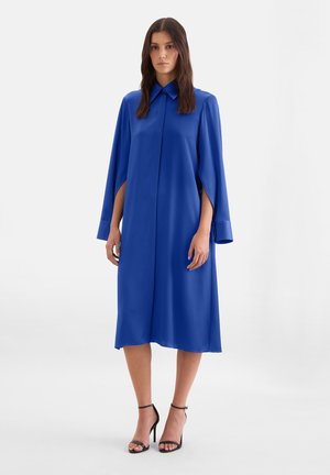 Femme portant une robe bleu roi longueur genoux avec des manches style cape et des sandales à talons hauts noires, debout devant un fond uni.