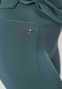 Zbliżenie na turkusowe legginsy sportowe z widocznymi szwami i małym trójkątnym logo "Kombat System Pro" na biodrze.