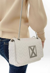 Borsa a tracolla bianca trapuntata con una catena argentata e un emblema geometrico. Tessuto liscio e forma strutturata. Dettagli accentuati comprendono componenti metallici.