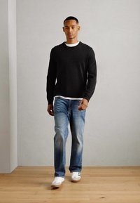 Schwarzer gerippter Langarmpullover über einem weißen T-Shirt, kombiniert mit hellblauen Jeans und weißen Sneakers, auf einem Holzboden.