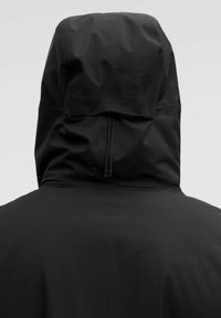 Sort vandtæt jakke med justerbar hætte set fra ryggen på en ensfarvet lys baggrund.