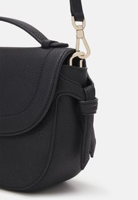 Musta teksturoidusta nahasta valmistettu laukku, jossa on kaartuva läppä, säädettävä hihna ja kullanväriset metallikoristeet sivuilla. Kompakti ja jäsennelty muotoilu.