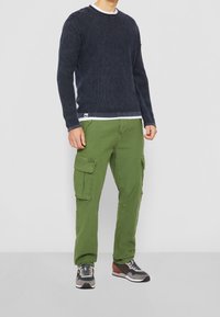 Marineblauer Strickpullover mit Rundhalsausschnitt, über einem weißen Hemd getragen, kombiniert mit olivgrünen Cargo-Hosen mit Seitentaschen und dunklen Sneakers.