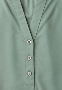 Devant de chemise vert clair avec col en V et trois petits boutons ronds alignés verticalement au centre.