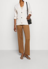 Kobieta ubrana w kremowy kardigan, brązowe szerokie spodnie w prążki, sandały na obcasie w wzór zebry, trzymająca czarną torebkę na ramię na białym tle.