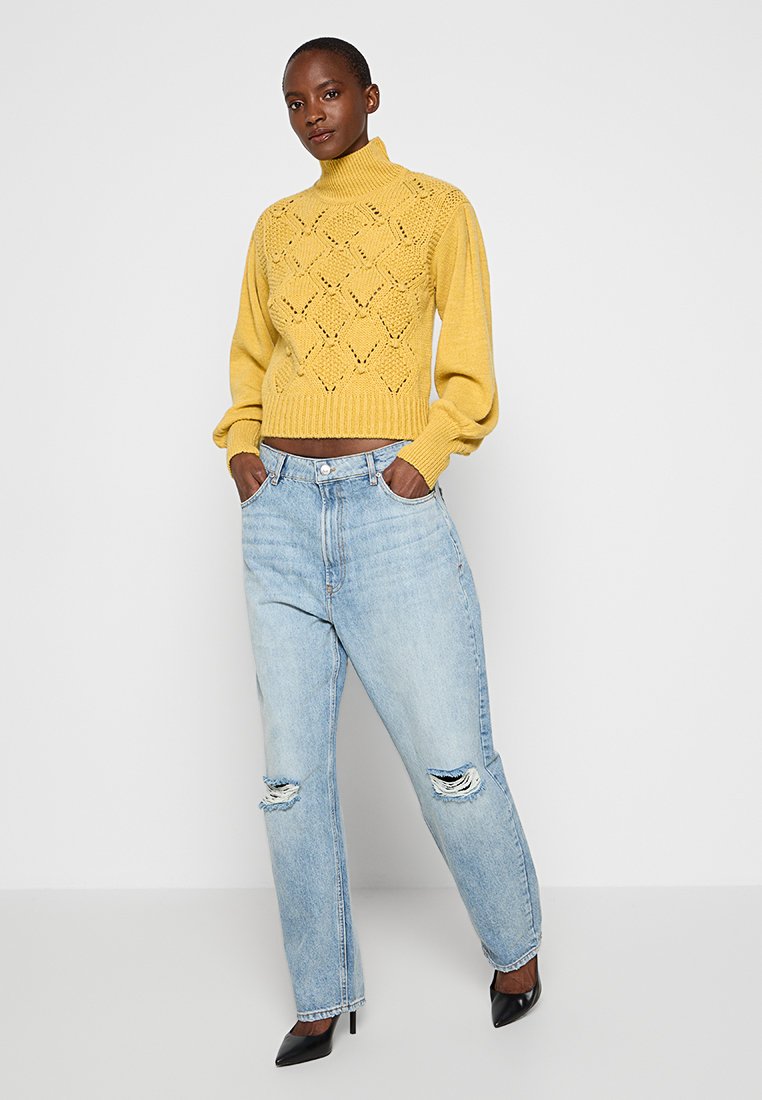 Donna in piedi con le mani nelle tasche, indossa un maglione a collo alto giallo lavorato a maglia, jeans strappati blu chiaro e décolleté neri.