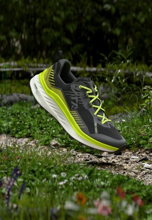 Scarpa da trail running nera e verde neon che fluttua sopra un sentiero roccioso e erboso con fiori selvatici e fogliame verde sullo sfondo.