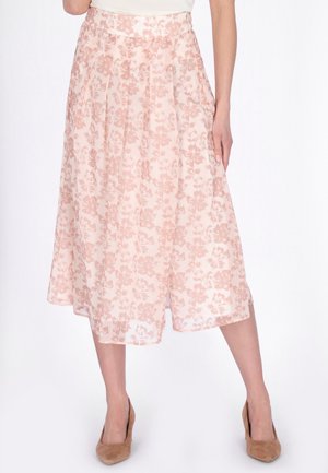 Spódnica midi w jasnoróżowy kwiatowy wzór, z bocznymi kieszeniami, noszona z cielistymi szpilkami z zaostrzonym noskiem oraz białą włożoną do środka bluzką, nogi lekko rozstawione.
