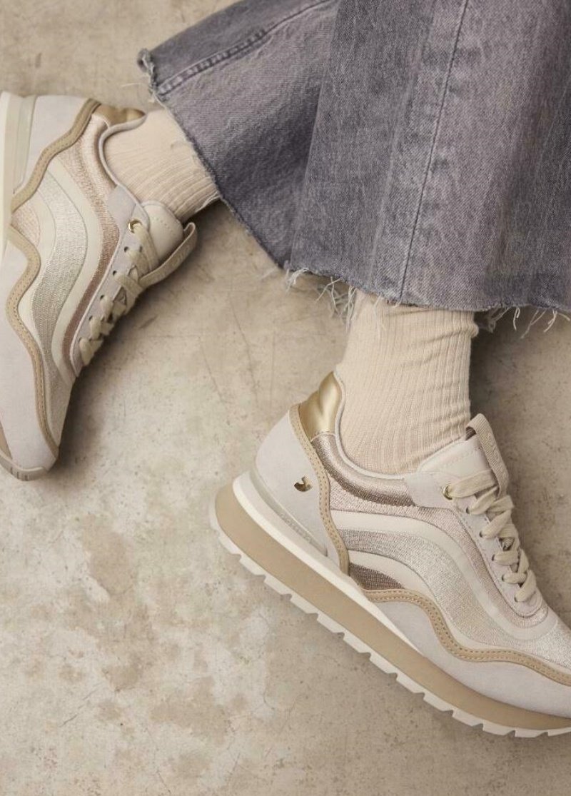 Chaussures de sport beiges avec un dessus texturé, des accents dorés et une semelle extérieure blanche. Associées à des chaussettes côtelées beige clair et à un pantalon en jean gris.