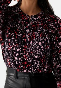 Blusa nera con un motivo sparso rosso e rosa, colletto con bottoni e dettagli plissettati. Tessuto morbido; abbinata a una cintura in pelle nera.