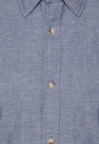 Camicia button-up in lino azzurro chiaro con colletto a punte, bottoni perlacei e una sottile texture con cuciture visibili lungo la patta.