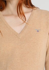 Beżowy sweter w serek noszony przez osobę z czerwonymi włosami, z małym wyszywanym logo GANT na piersi.