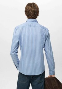 Svetlo modra dolga srajca z belimi navpičnimi črtami, gumbi na ovratniku in zaokroženim robom. Model nosi temne jeans hlače in drži rjavo jakno.