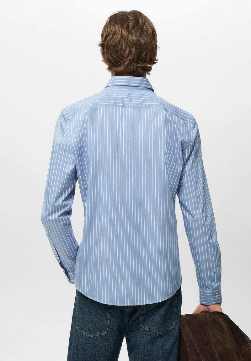 Hellblaues Langarmhemd mit weißen vertikalen Streifen, durchgeknöpfter Kragen und abgerundetem Saum. Das Modell trägt dunkle Jeans und hält eine braune Jacke.