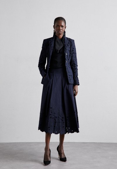 Navy lace blazer with floral pattern, paired with a pleated navy skirt featuring a detailed hem. Black high-heeled shoes complete the look.