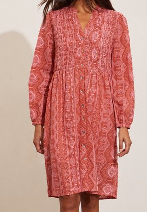 Czerwona sukienka w geometryczne wzory, z długimi rękawami, marszczonym gorsetem, zapięciem na guziki z przodu i skomplikowanymi geometrycznymi wzorami na całej powierzchni.