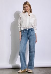 Bílá košile na knoflíky s náprsní kapsou, světle modré široké džíny a béžové sandály s fleece podšívkou a texturovanými detaily.