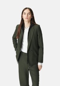 Giacca blazer verde oliva con un design strutturato, due bottoni frontali e rever affilati. Abbinata a una camicia bianca e a un giubbotto nero con cappuccio.