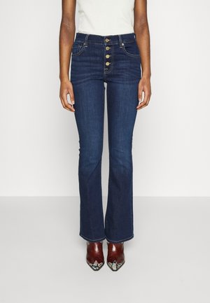 Osoba ubrana w granatowe, wysokie dzwony z czterema widocznymi złotymi guzikami oraz brązowe buty na szpic z kolorowymi detalami.